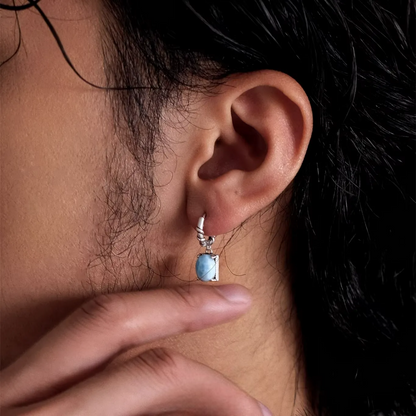 Silver huggie earring with a light blue larimar stone drop on an ear.